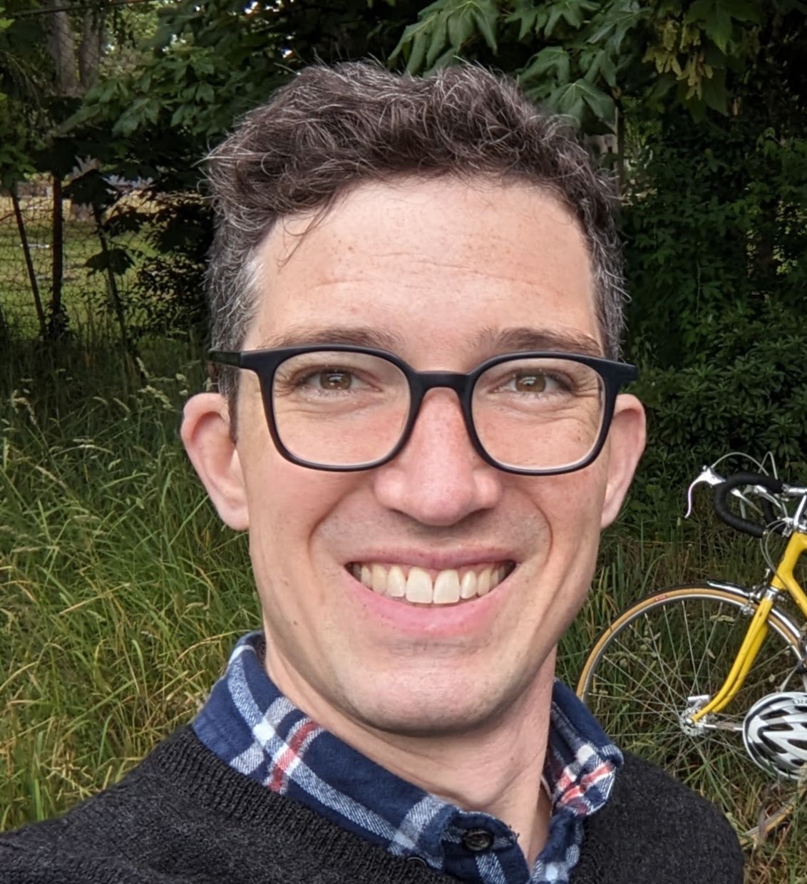 Peter in a flannel shirt and sweater, in front of long green grass, some green trees, and a bright yellow Schwinn super sport bicycle.