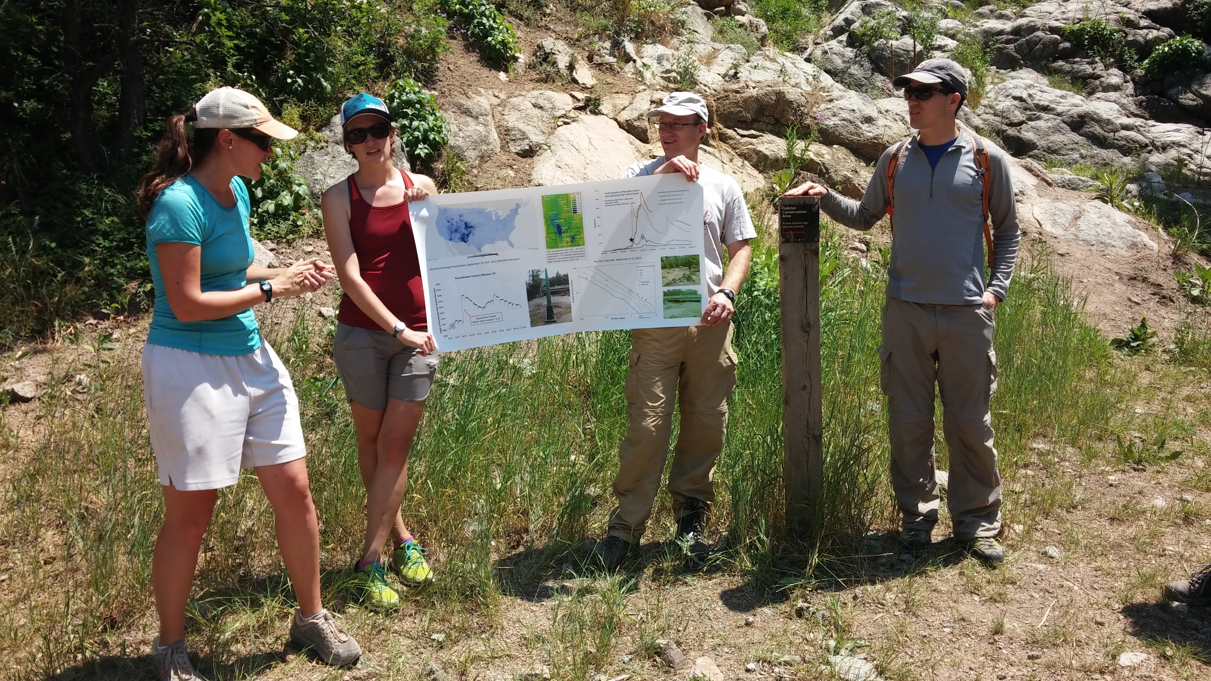 Four people standing in front of shrubs and rocks in Boulder, Colorado, 
                                      two of whom are holding
                                      a poster. One of the people is describing the information on the poster to 
                                      the viewer.