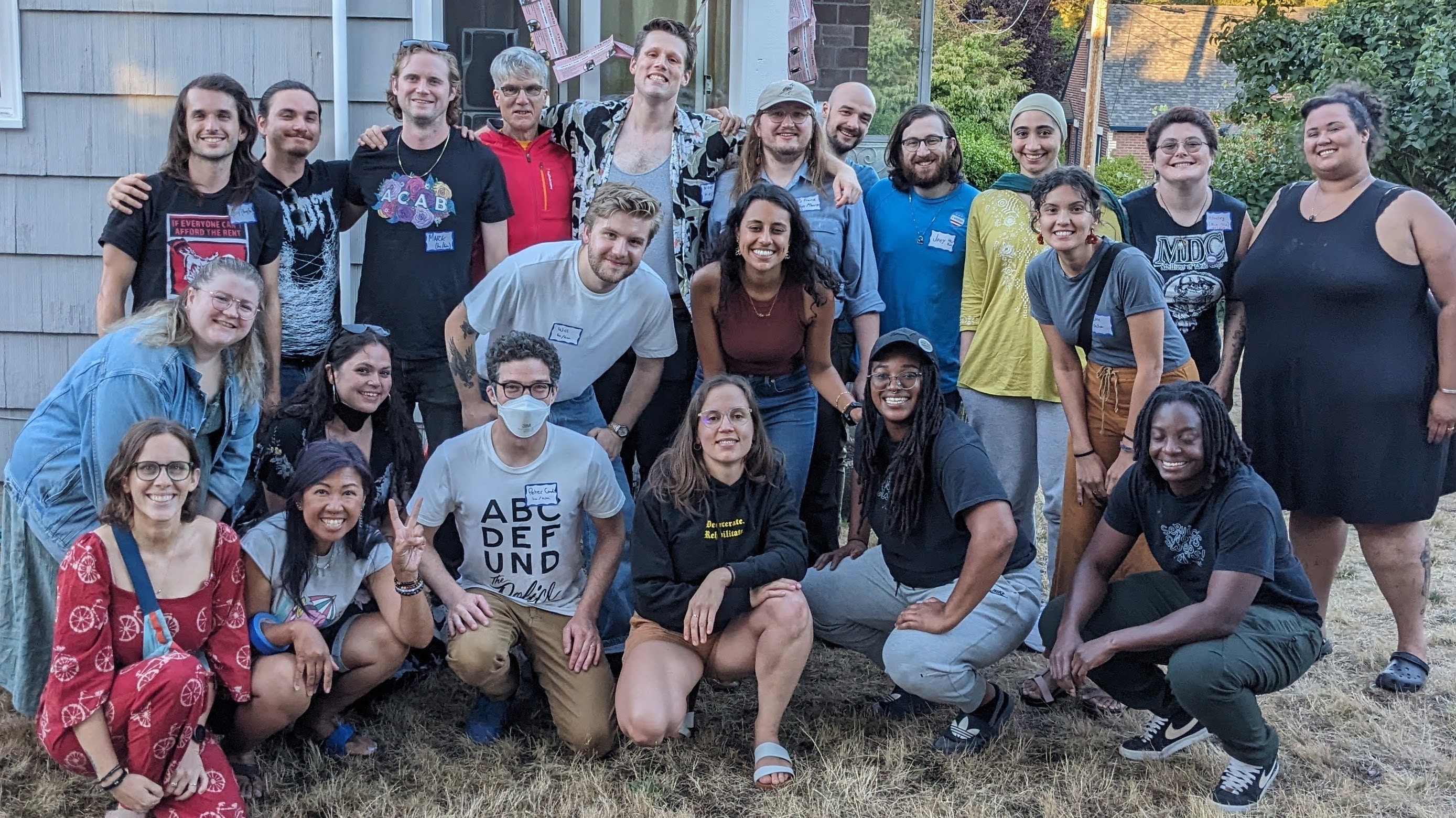 22 people in casual clothing posing in a yard in front of a small house. Many have arms around 
                                each other and some are kneeling in front.